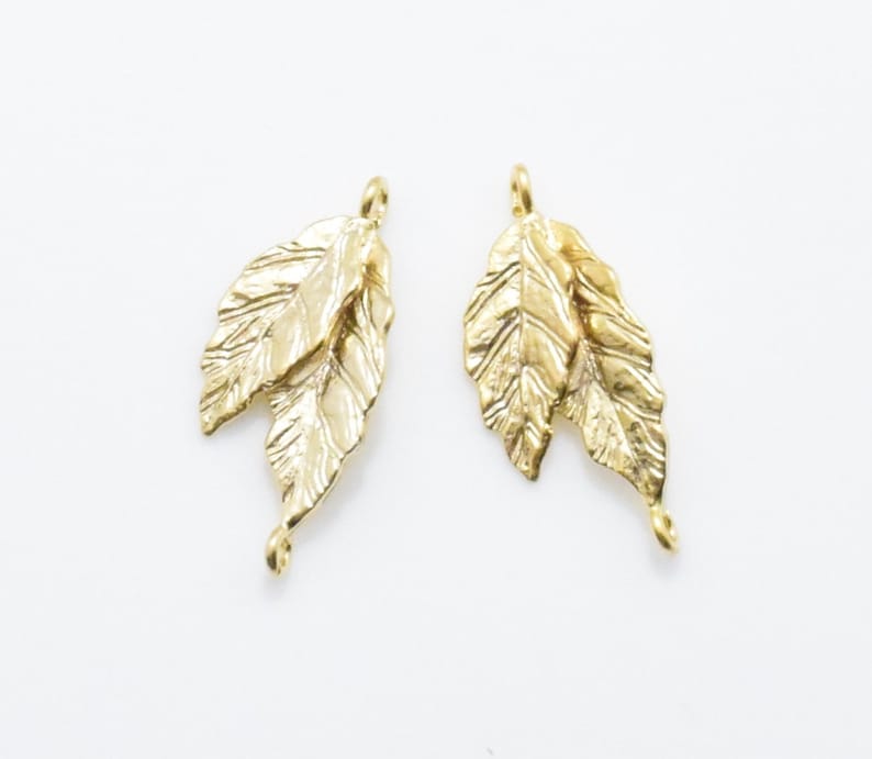 Pode incluir: Dois pingentes de metal dourado em forma de folha com uma superf&iacute;cie texturizada. Cada pingente tem duas folhas e um la&ccedil;o na parte superior para prender em joias.