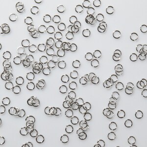 Puede incluir: Un conjunto de anillos de salto plateados sobre un fondo blanco. Los anillos de salto son pequeños y redondos, y son perfectos para la elaboración de joyas.