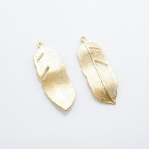 May include: Two gold-colored metal feather charms with a textured surface. The feathers have a hole at the top for attaching to jewelry or other crafts.