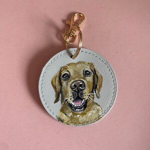 May include: A round, white canvas keychain with a gold metal clasp. A hand-painted portrait of a golden retriever dog with brown eyes and a happy expression is featured on the keychain.