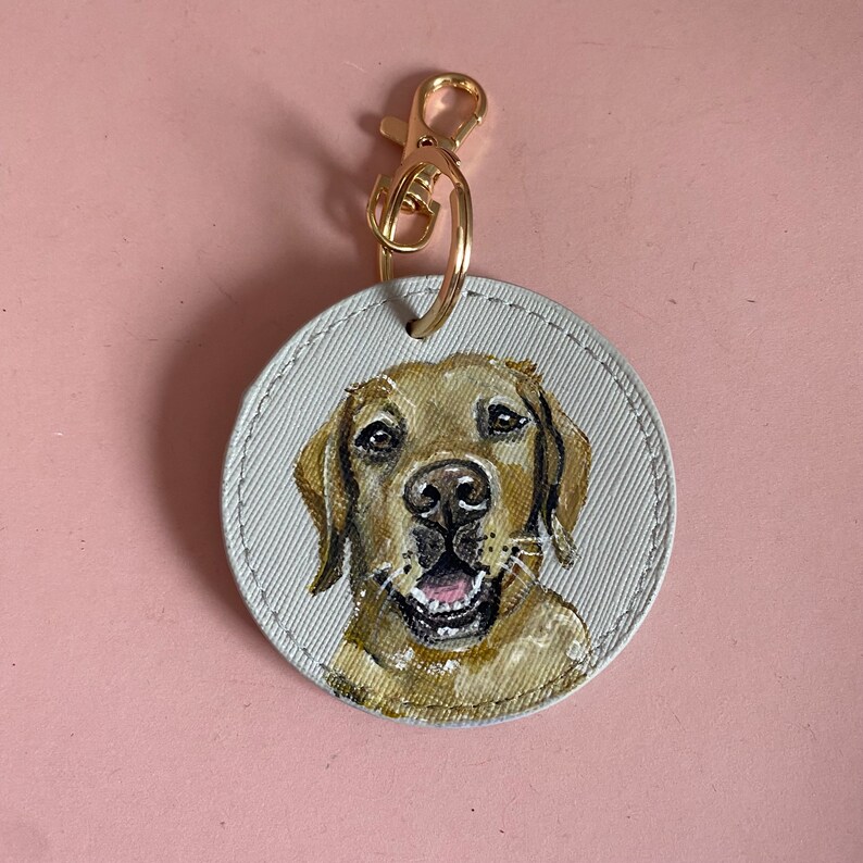 May include: A round, white canvas keychain with a gold metal clasp. A hand-painted portrait of a golden retriever dog with brown eyes and a happy expression is featured on the keychain.