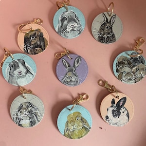May include: Nine round keychains with painted images of rabbits. The keychains are made of leather and have a gold metal ring. The rabbits are painted in various colors, including brown, black, white, and gray.