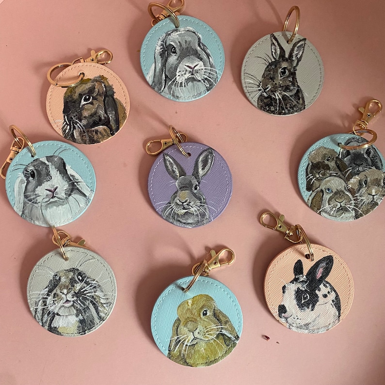 May include: Nine round keychains with painted images of rabbits. The keychains are made of leather and have a gold metal ring. The rabbits are painted in various colors, including brown, black, white, and gray.