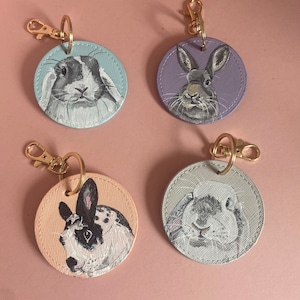 May include: Four round keychains with painted images of rabbits. Each keychain is a different color: light blue, purple, light pink, and light gray. The rabbits are all different breeds and have unique markings.