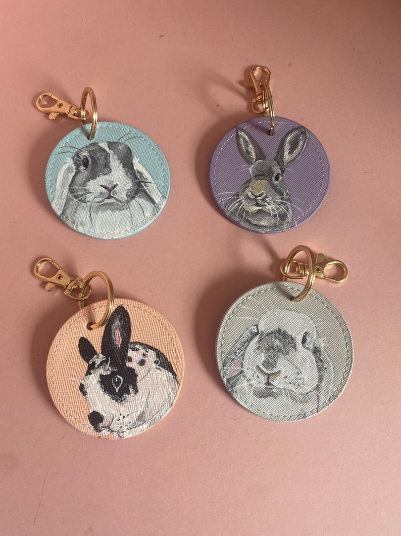 May include: Four round keychains with painted images of rabbits. Each keychain is a different color: light blue, purple, light pink, and light gray. The rabbits are all different breeds and have unique markings.