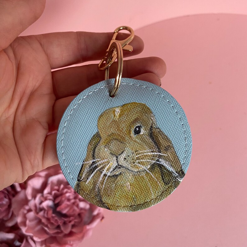 May include: A round, light blue keychain with a stitched edge features a hand-painted portrait of a brown rabbit with white whiskers. The keychain has a gold metal ring and clasp.