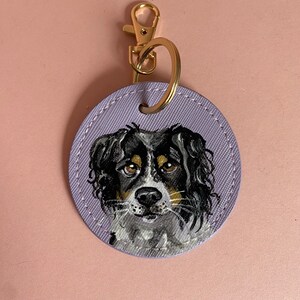 May include: A purple leather keychain with a painted portrait of a black and white dog with brown eyes. The dog is looking directly at the viewer.