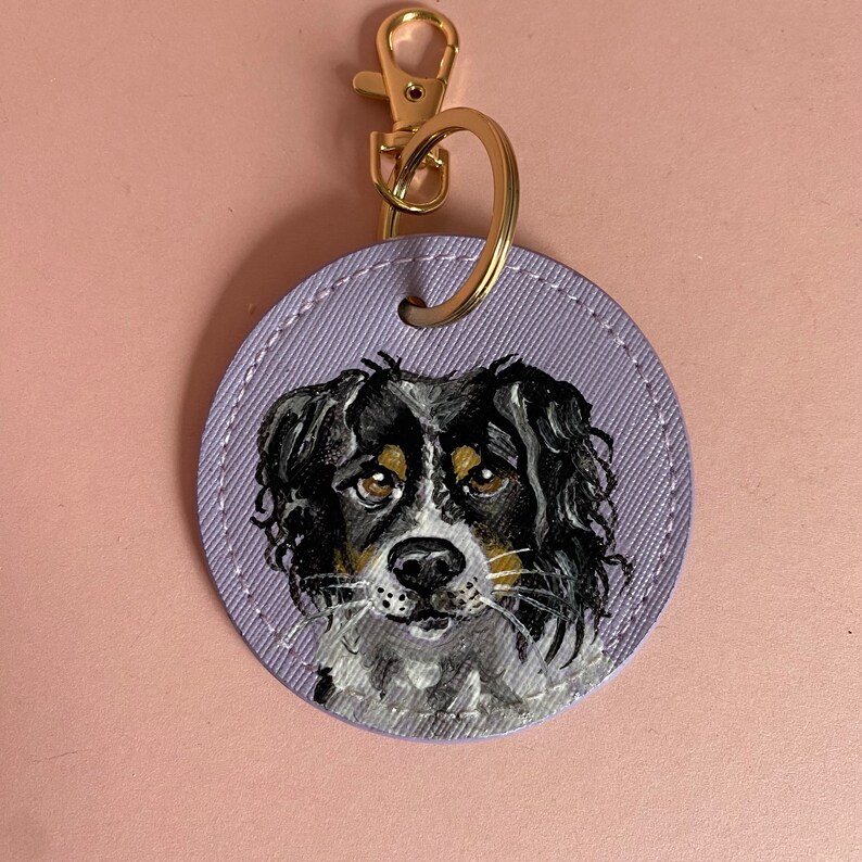 May include: A purple leather keychain with a painted portrait of a black and white dog with brown eyes. The dog is looking directly at the viewer.