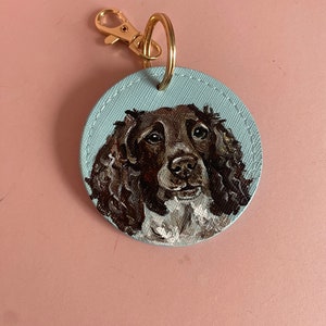May include: A round, blue leather keychain with a painted portrait of a brown and white dog. The dog has brown eyes and a white patch on its chest.