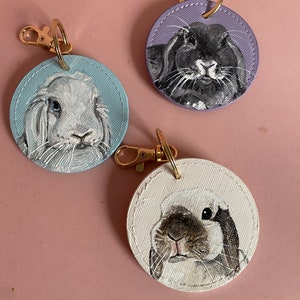 May include: Three round leather keychains with painted portraits of rabbits. One keychain features a black rabbit on a purple background, another features a white rabbit on a blue background, and the third features a brown and white rabbit on a white background.