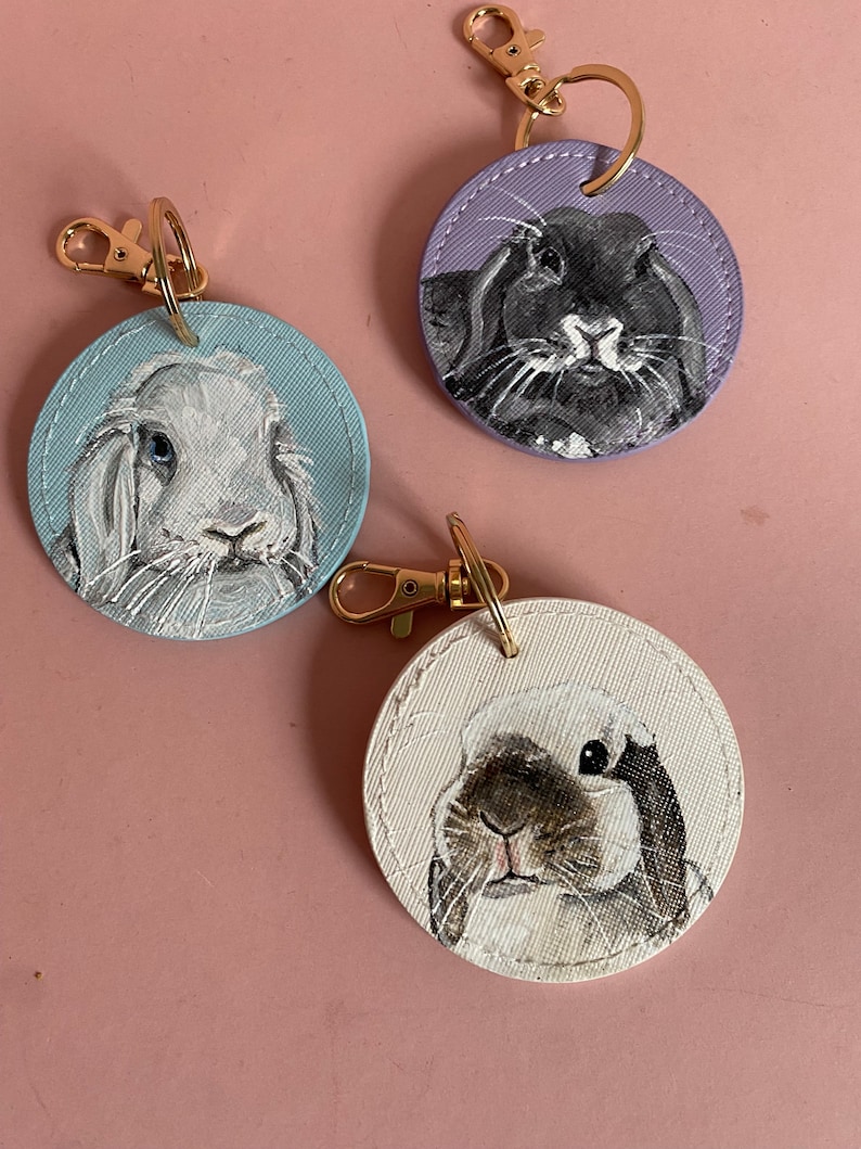 May include: Three round leather keychains with painted portraits of rabbits. One keychain features a black rabbit on a purple background, another features a white rabbit on a blue background, and the third features a brown and white rabbit on a white background.