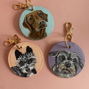 May include: Three round leather keychains with painted portraits of dogs. The first keychain features a brown dog with a light blue background. The second keychain features a black and white dog with a light pink background. The third keychain features a gray dog with a light purple background.
