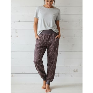 Op de afbeelding: Een persoon draagt een lichtgrijs T-shirt met ronde hals en een donkerbruine joggingbroek met patroon en zakken. De joggingbroek heeft een donkergrijze tailleband en manchetten. De achtergrond is een witte houten muur.