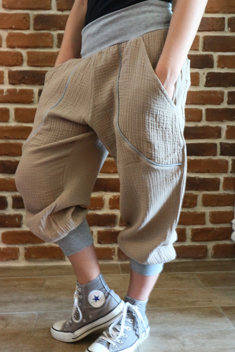 May include: A pair of beige, loose-fitting pants with a gray waistband and contrasting stitching. The pants have pockets on the sides and are cuffed at the ankles. The person is wearing gray high-top sneakers.