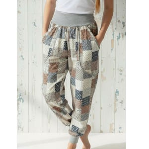 Könnte beinhalten: Haremshose mit Patchworkmuster in Beige, Braun und Marineblau. Die Hose hat einen grauen Bund und Bündchen. Das Model trägt ein weißes Oberteil und hat die Hände in den Hosentaschen.