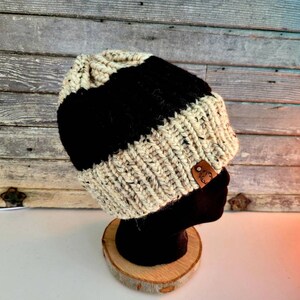 May include: A black and white knitted beanie with a brown leather patch. The beanie is on a wooden stand.