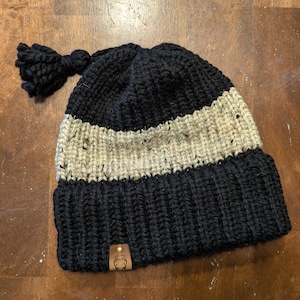 May include: Black and beige knitted beanie with a black pom-pom on top. The beanie has a brown leather patch with a white logo on the side.