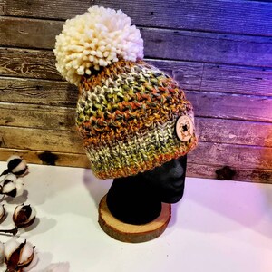 May include: A multi-colored knitted beanie with a large white pom pom on top. The beanie has a brown button with a design on it.