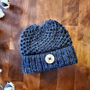May include: A dark blue crocheted beanie with a brown button featuring a bear design. The button has the text "Good Vibes" on it.
