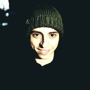 May include: A close-up portrait of a person wearing a dark green knitted beanie. The person is smiling and looking directly at the camera.