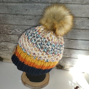 May include: A knitted hat with a large faux fur pom-pom. The hat has a colour block design with navy blue, yellow, and white sections. The top of the hat has a speckled pattern with various colours. A wooden button is on the side.