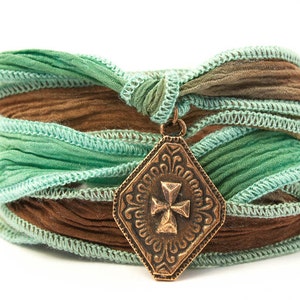 May include: A multi-layered bracelet made of brown and green fabric with a decorative metal pendant featuring a cross design.