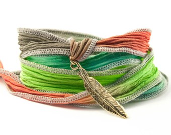 Könnte beinhalten: Ein mehrfarbiges Seidenband-Armband mit einem goldenen Federschmuck. Das Armband besteht aus mehreren Schichten Band in Grün-, Braun- und Orangetönen.