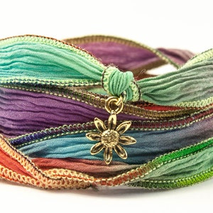 May include: Multicoloured fabric bracelet with a gold sunflower charm. The bracelet is made of several layers of fabric, each with a different colour and pattern. The sunflower charm is attached to the bracelet with a gold loop.