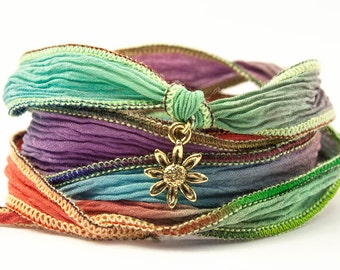 Könnte beinhalten: Mehrfarbiges Stoffarmband mit einem goldenen Sonnenblumen-Anhänger. Das Armband besteht aus mehreren Stofflagen, jede mit einer anderen Farbe und einem anderen Muster. Der Sonnenblumen-Anhänger ist mit einer goldenen Schlaufe am Armband befestigt.