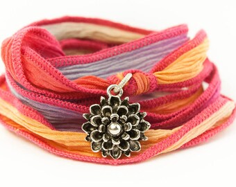 Könnte beinhalten: Ein mehrfarbiges Seidenband-Armband mit einem silbernen Blumenanhänger. Das Armband besteht aus mehreren Schichten Band in den Farben Rosa, Lila und Orange.