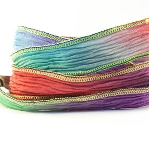 May include: A roll of colorful, pleated ribbon in shades of purple, red, green, and blue. The edges are trimmed with a contrasting gold and green stitching, creating a decorative accent. The ribbon is likely used for crafts or gift wrapping.