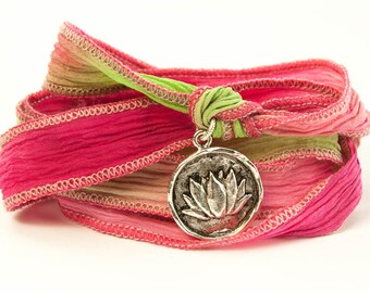Könnte beinhalten: Ein Armband aus rosa, grünem und beigefarbenem Seidenband mit einem silbernen Lotusblüten-Anhänger.
