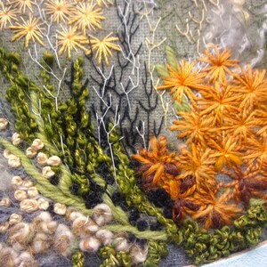 A detailed macro close-up of a hand-embroidered floral meadow study showcasing dense orange and yellow wool yarn sunburst blooms, fine white and green branching stitches, and tightly clustered French knots on a textured linen background, framed by a light blue oval mount.