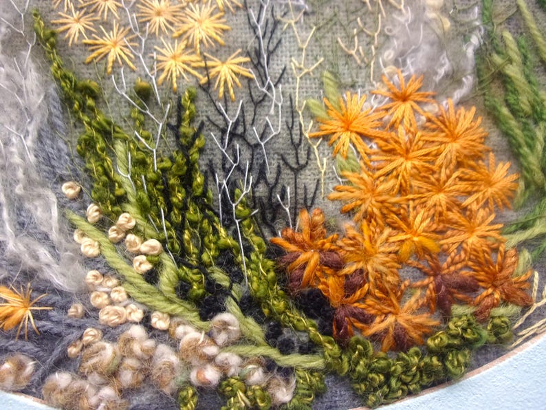 A detailed macro close-up of a hand-embroidered floral meadow study showcasing dense orange and yellow wool yarn sunburst blooms, fine white and green branching stitches, and tightly clustered French knots on a textured linen background, framed by a light blue oval mount.