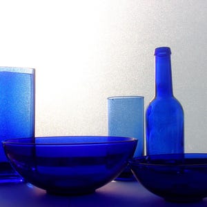 Cluster of blue glassware featuring cobalt blue bowls, tumblers, and a tall vintage cobalt blue bottle, arranged together against a light neutral background.
