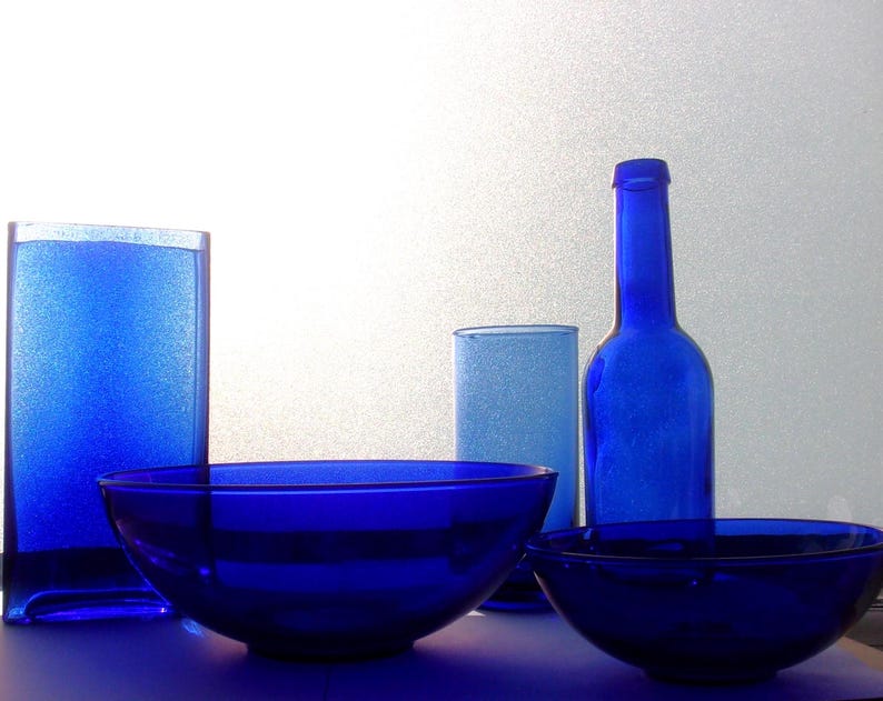 Cluster of blue glassware featuring cobalt blue bowls, tumblers, and a tall vintage cobalt blue bottle, arranged together against a light neutral background.