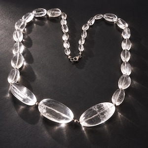 Pode incluir: Um colar composto por contas de cristal transparentes, polidas e de formato oval, de tamanhos variados. As contas estão enfiadas com pequenos espaçadores metálicos entre elas, e o colar é preso com um fecho de mosquetão de metal prateado.