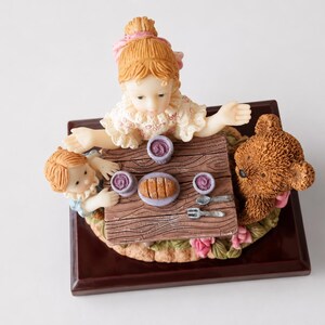 May include: A decorative figurine featuring two children and a brown teddy bear sitting at a small wooden table. The table is set with three purple bowls, a loaf of bread on a plate, a silver spoon, and a silver fork, all mounted on a dark base.