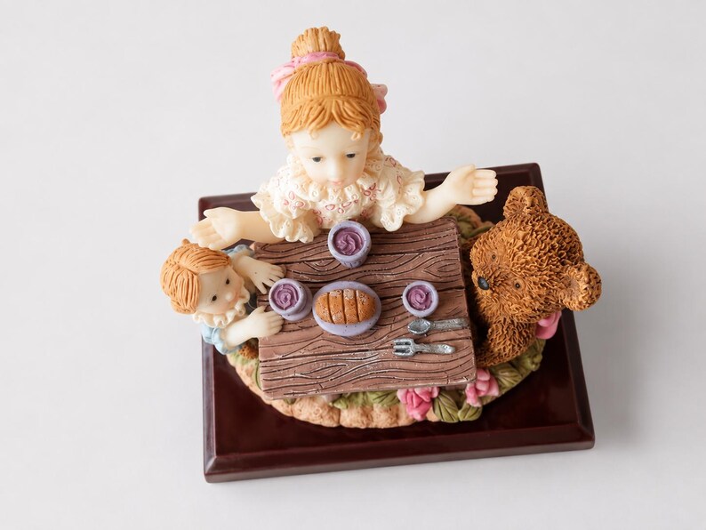 May include: A decorative figurine featuring two children and a brown teddy bear sitting at a small wooden table. The table is set with three purple bowls, a loaf of bread on a plate, a silver spoon, and a silver fork, all mounted on a dark base.