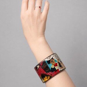 Handmade Comic Book Pop Art bangle worn on a hand, showing a wide glossy upcycled wood cuff covered in colourful vintage comic collage artwork with illustrated faces, speech bubbles, red and pink graphic details, and bold comic imagery. Shown against a clean light-grey studio background.