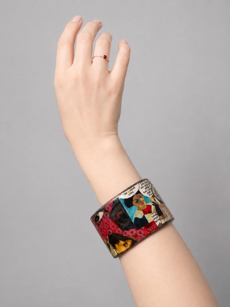Handmade Comic Book Pop Art bangle worn on a hand, showing a wide glossy upcycled wood cuff covered in colourful vintage comic collage artwork with illustrated faces, speech bubbles, red and pink graphic details, and bold comic imagery. Shown against a clean light-grey studio background.