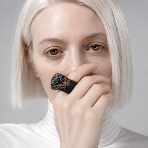 May include: A close-up of a black ring with a rough, textured surface. The ring is held up to the face, with the focus on the ring's unique design and the contrast against the skin and white turtleneck.