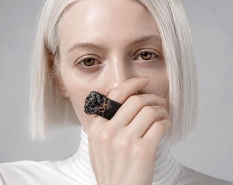 May include: A close-up of a black ring with a rough, textured surface. The ring is held up to the face, with the focus on the ring's unique design and the contrast against the skin and white turtleneck.