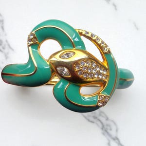 May include: A gold-toned snake-shaped bracelet with a turquoise enamel finish. The snake's head and details are encrusted with clear crystals. The bracelet has a hinged design and is set against a white marble background.