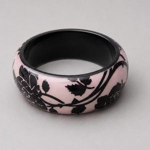 May include: A wide bangle bracelet featuring a smooth, pale pink base with a contrasting black floral pattern of roses and leaves wrapping around the exterior. The interior of the circular bracelet is solid black, providing a distinct color contrast.