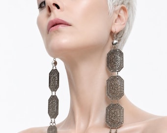 May include: A pair of long, dangling earrings featuring three vertically stacked, octagonal metal plates with intricate filigree patterns. Each earring is finished with two small, round, textured coin-like charms at the bottom, suspended from a silver-toned hook.