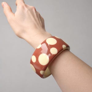 May include: A wide, glossy bangle bracelet featuring a terracotta-colored base with a pattern of cream-colored polka dots of varying sizes. The bracelet has a smooth, rounded surface and a thick, substantial profile that encircles the wrist.