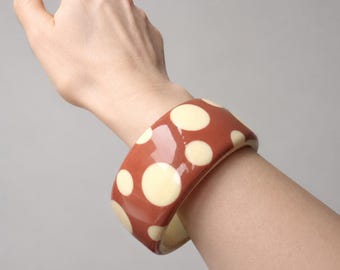 May include: A wide, glossy bangle bracelet featuring a terracotta-colored base with a pattern of cream-colored polka dots of varying sizes. The bracelet has a smooth, rounded surface and a thick, substantial profile that encircles the wrist.
