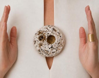 May include: A round, speckled brooch with a textured surface, featuring a mix of white, black, and brown tones. The brooch is set against a white garment, with a gold ring on the right hand.