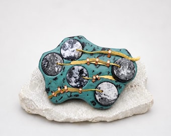May include: A unique, abstract brooch with a teal base and four circular inlays of black and white stone. The brooch is accented with gold and copper-colored metal details, and rests on a white stone base. This handcrafted jewelry piece is a wearable art statement.